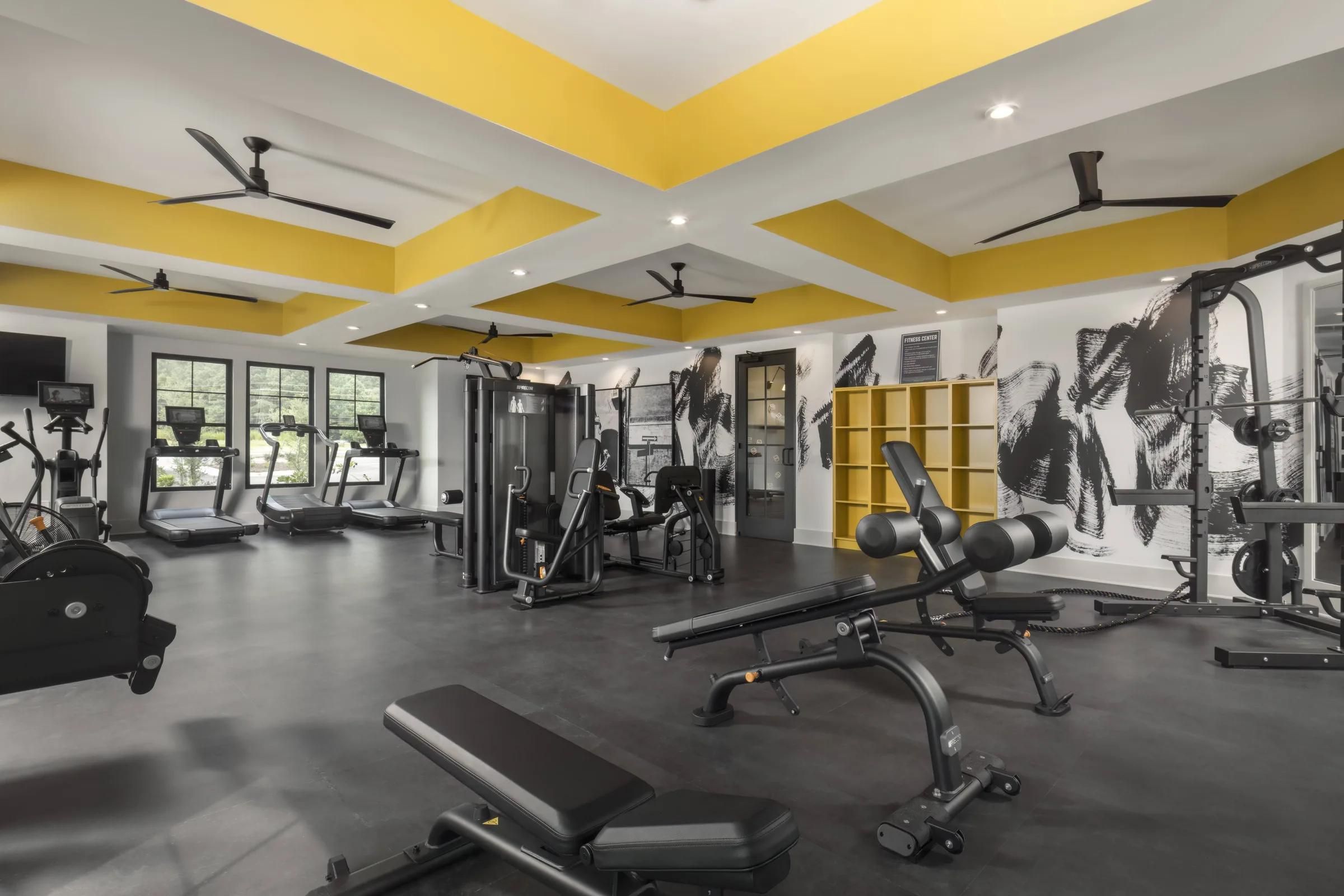 Evolve at the Pines modern gym with yellow ceiling accents, various exercise machines, benches, and wall art in black and white.