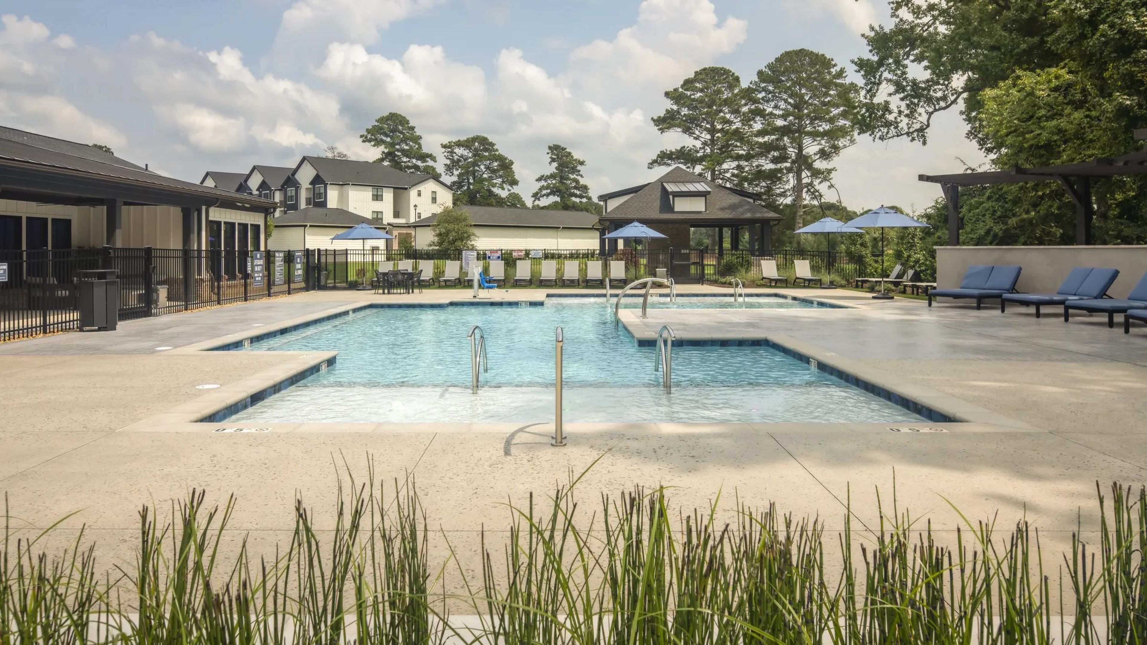 Evolve at the Pines outdoor swimming pool with lounge chairs and umbrellas, surrounded by buildings and tall trees under a cloudy sky.