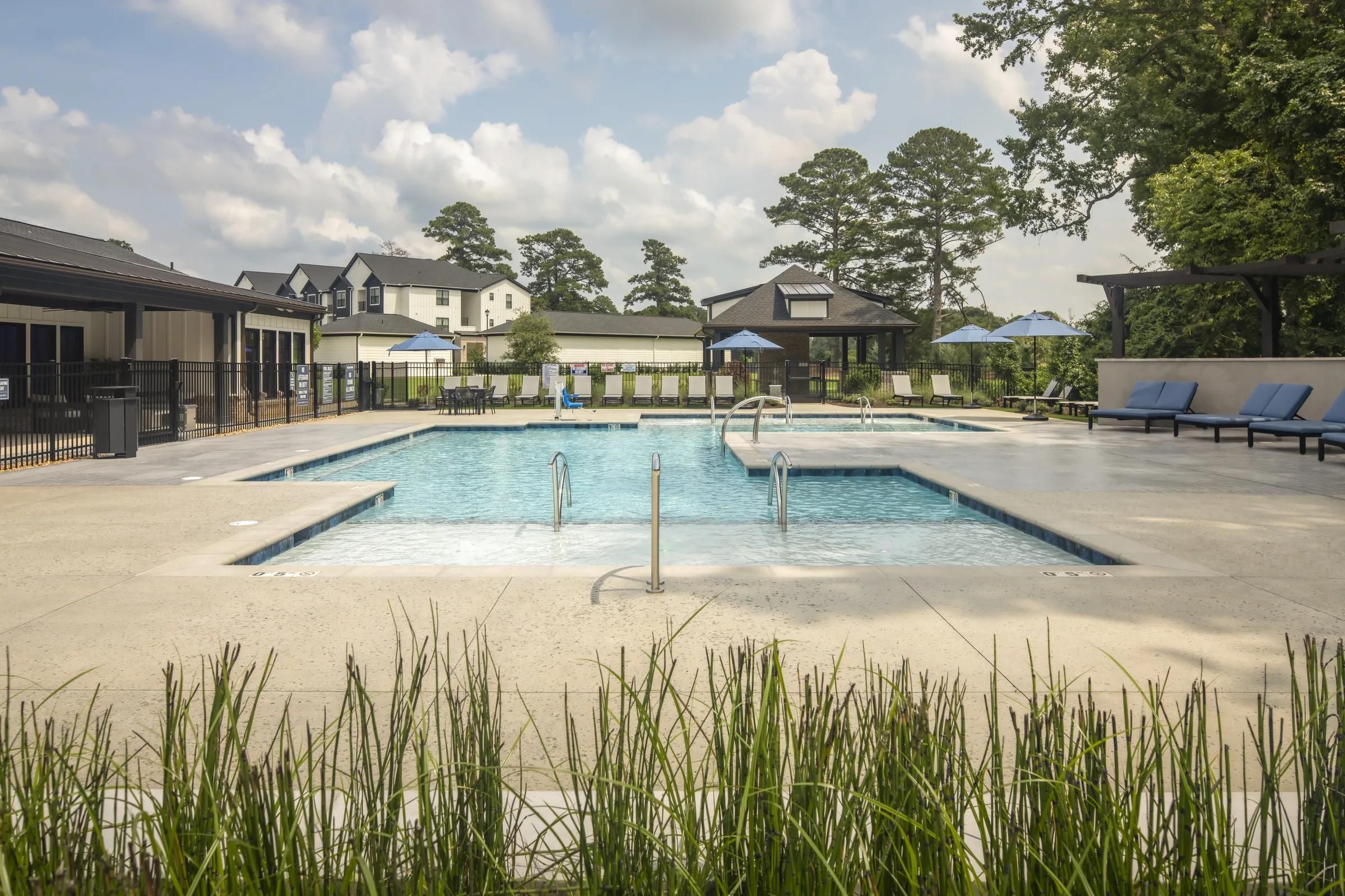 Evolve at the Pines outdoor swimming pool with lounge chairs and umbrellas, surrounded by buildings and tall trees under a cloudy sky.